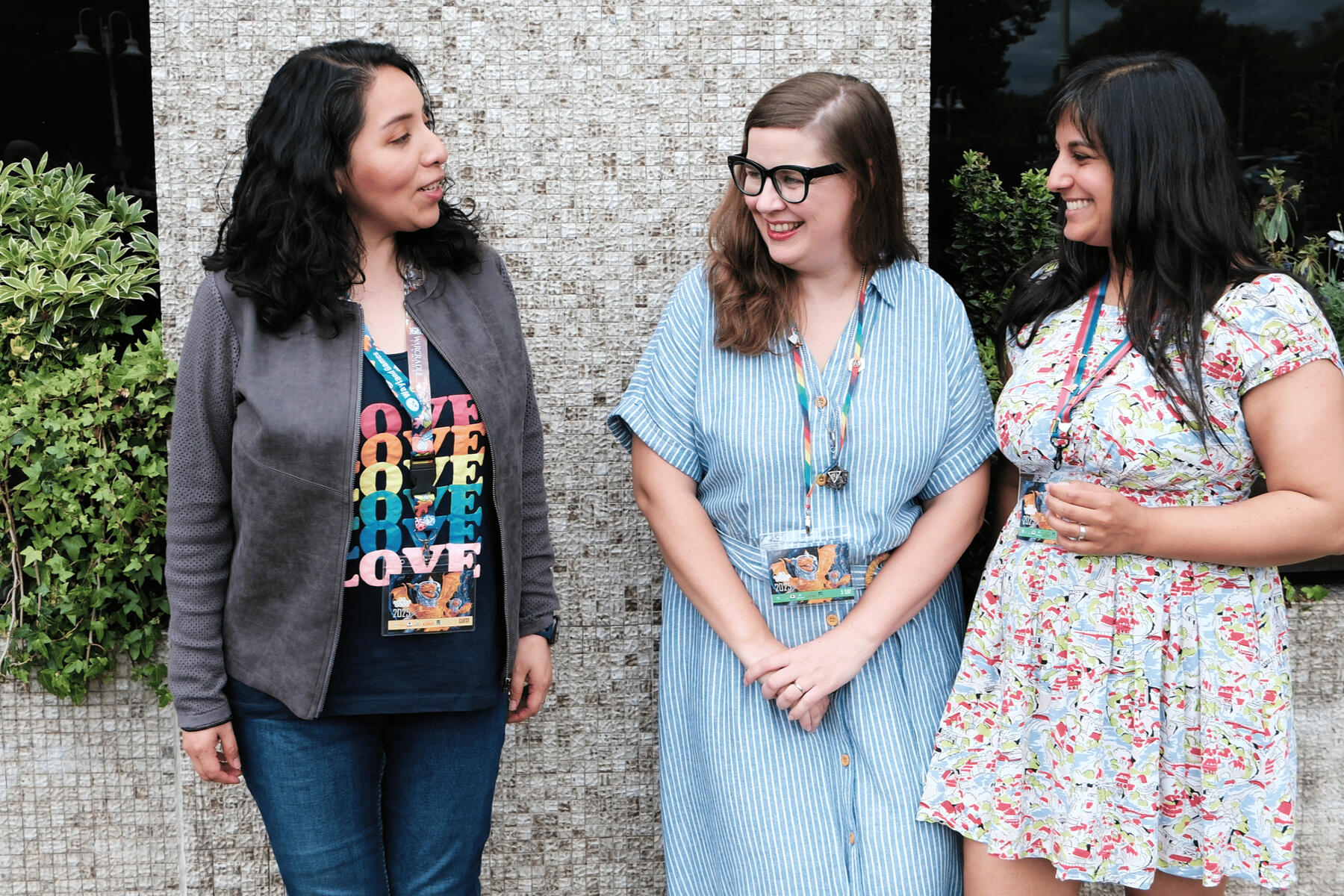 Three femme-presenting people standing talking and laughing together outdoors. Left to right, Aida, Darcy and Michelle